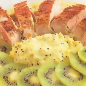 Plat de cuisine composé de tranches de viande de porc grillées accompagnées d'une purée onctueuse parsemée de graines de kiwi, entouré de tranches de kiwi frais disposées en éventail. Présentation soignée et colorée mettant en valeur les contrastes entre le vert vif des fruits et le doré de la viande